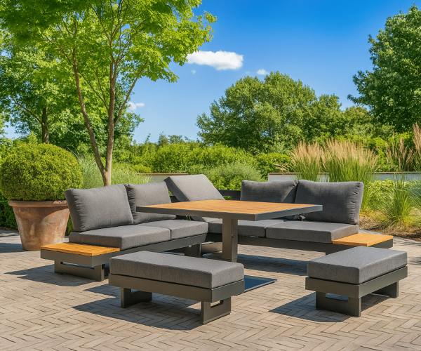 Aluminium-Gartenlounge Set mit dunkelgrauen Kissen, Teakholz-Tisch und zwei Hockern auf einer Pflastersteintetrasse in einem grünen Garten